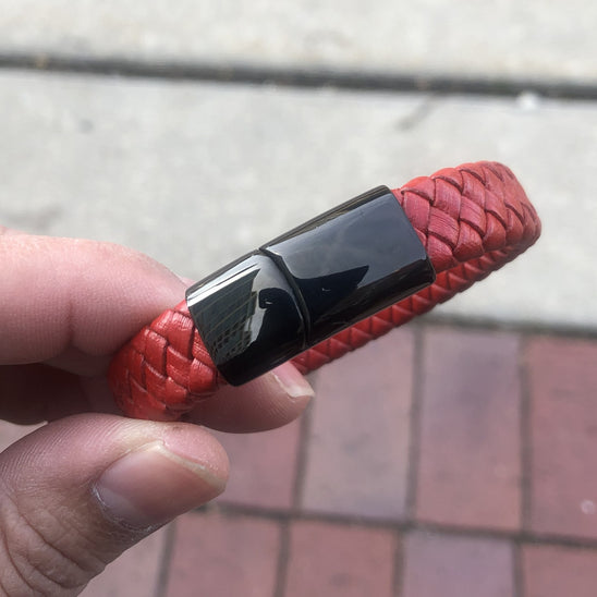 Red braided bracelet with a black clasp held by a hand on a blurred pavement background