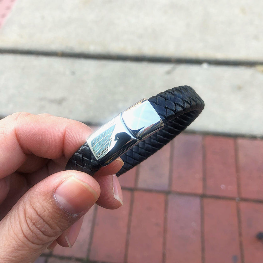 Black braided bracelet with a metallic clasp held by a hand on a concrete surface.