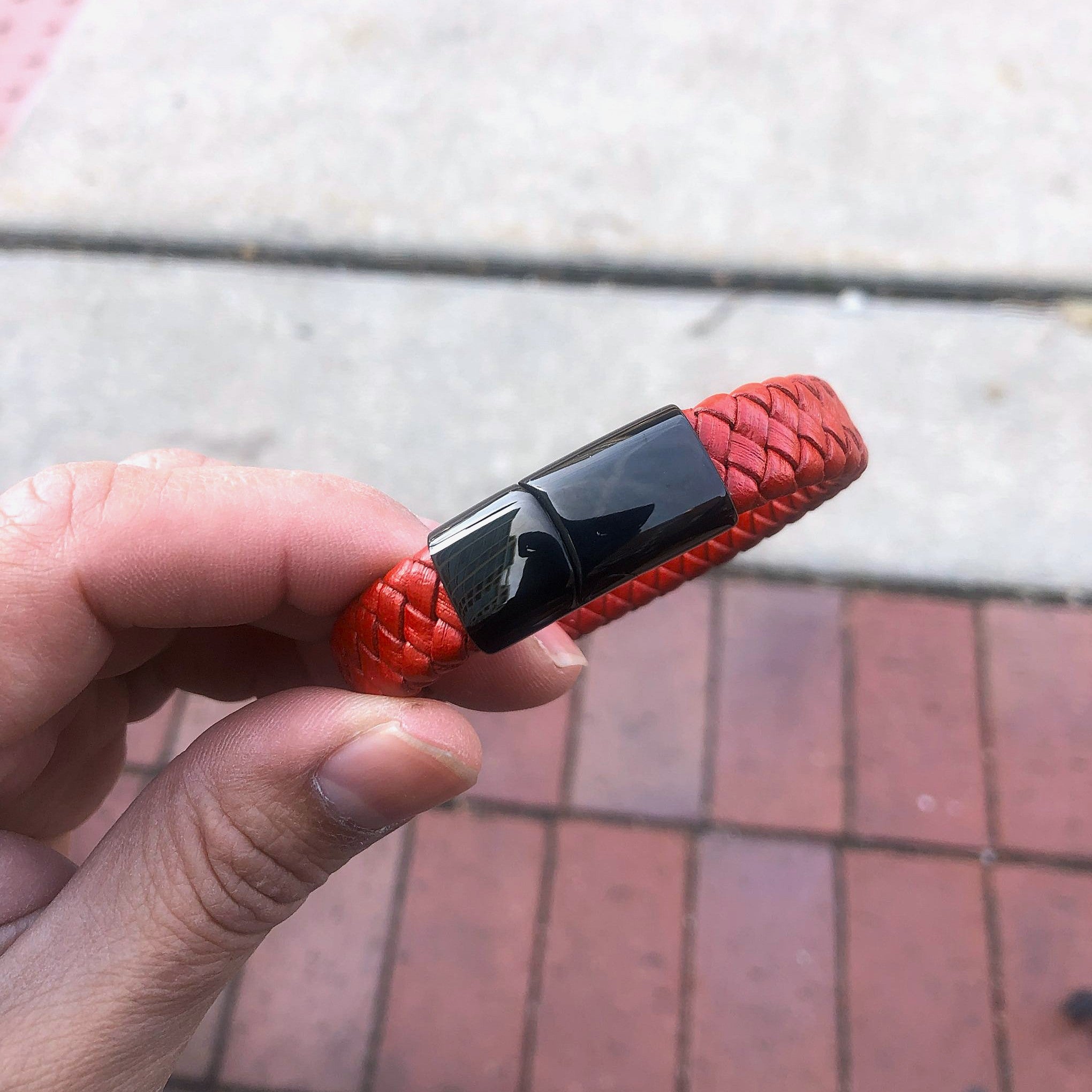 Red braided bracelet with a black clasp held by a hand on a pavement background