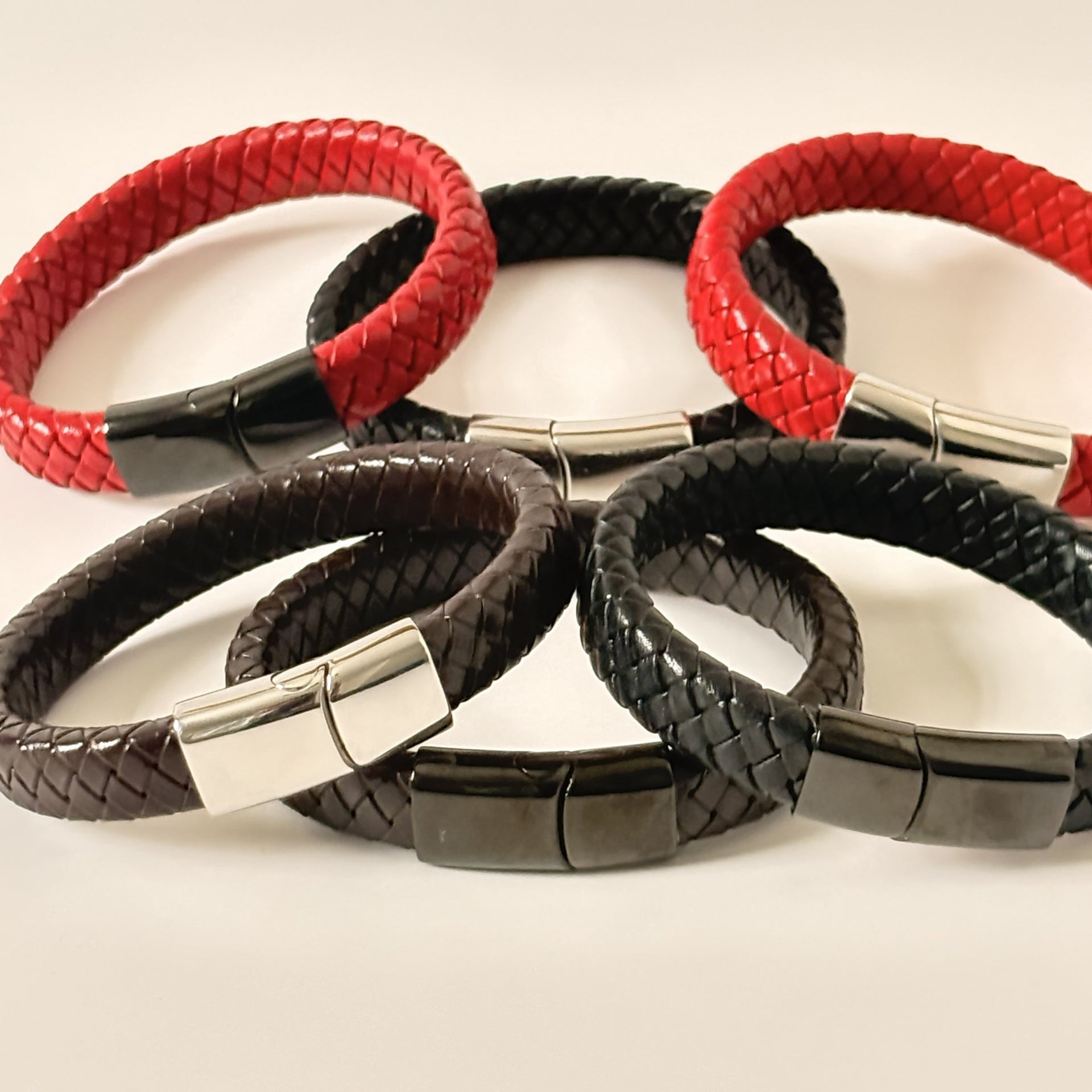 Four braided leather bracelets in red, black, and dark brown, each with a polished silver rectangular clasp, arranged on a light beige background.